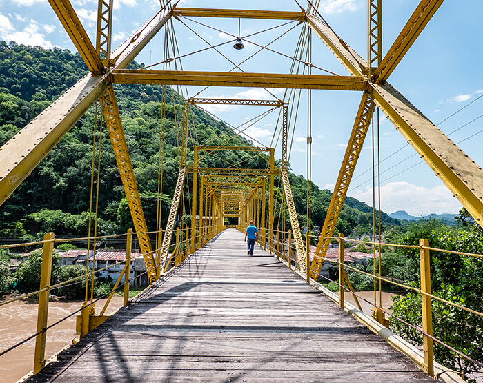 Puente metálico amarillo sobre un río en una zona montañosa, con una persona caminando por su estructura de madera.